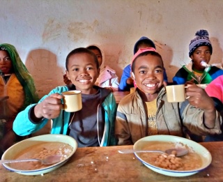 Two boys sit at a desk with bowls in front of them holding mugs up to the camera